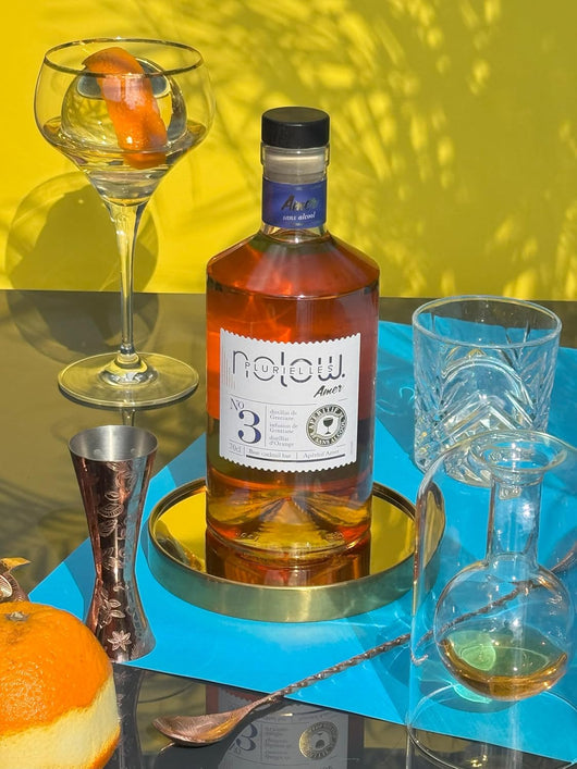Bottles of NOLOW N°3 Distillat Amer sans alcool with cocktail glasses and orange on a colorful table setting.